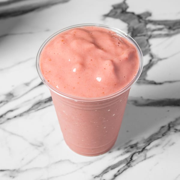 Pink strawberry smoothie in a clear plastic cup on a white marble surface.