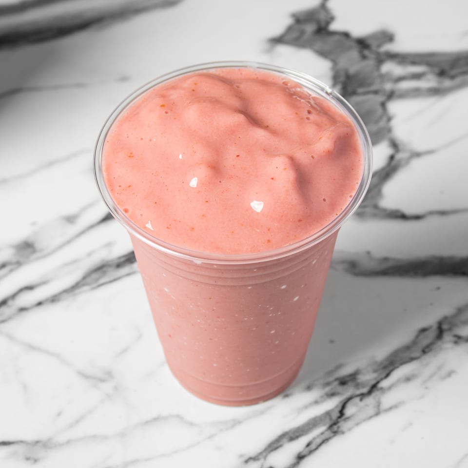 Pink strawberry smoothie in a clear plastic cup on a white marble surface.