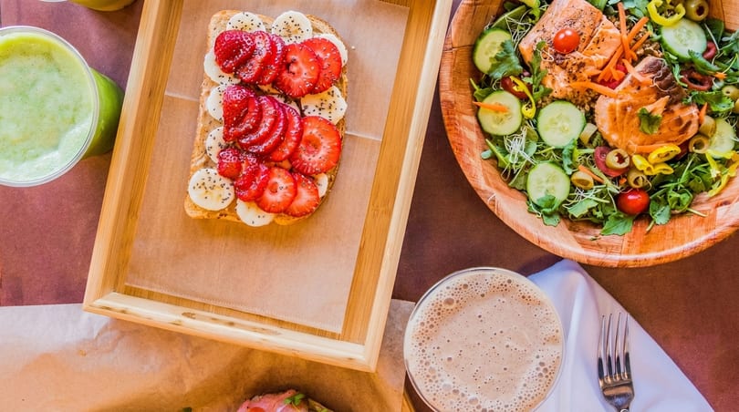 Overhead view of strawberry banana toast, salmon salad, green smoothie, and a chocolate drink.