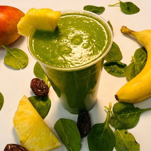 Green smoothie in a clear cup with pineapple, banana, apple, spinach, and dates.
