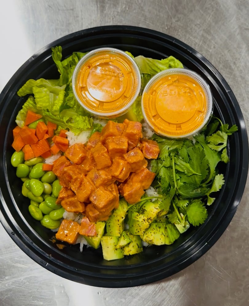 A poke bowl with salmon, avocado, edamame, carrots, cilantro, and two sauce cups.