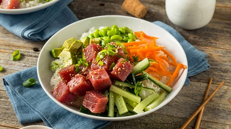 A colorful poke bowl with tuna, avocado, edamame, carrots, and cucumbers on rice.