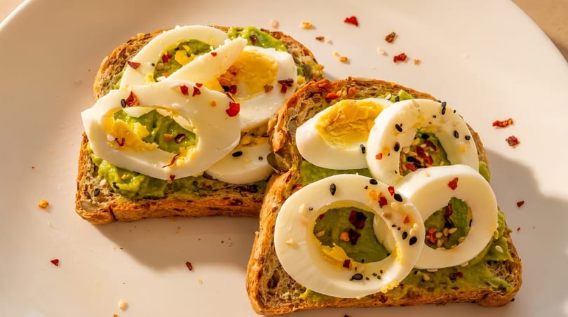 Two slices of avocado toast topped with sliced hard-boiled eggs and chili flakes on a white plate.