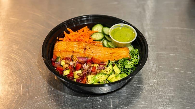 Salmon bowl with avocado, salsa, carrots, cucumbers, cilantro, and green dressing on a metal surface.