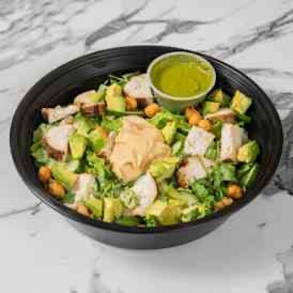 A black bowl of salad with chicken, avocado, chickpeas, lettuce, and a side of green dressing.