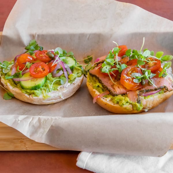 Two avocado toasts with various toppings, including tomatoes, onions, and microgreens, on parchment paper.
