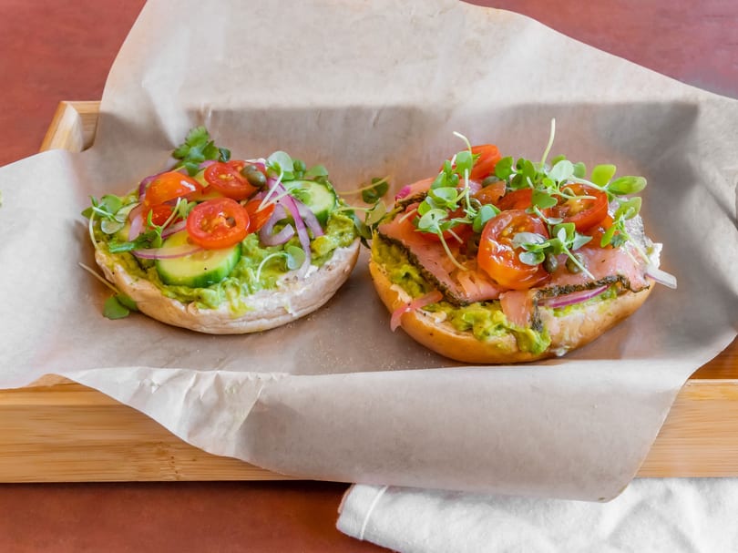 Two avocado toasts with various toppings, including tomatoes, onions, and microgreens, on parchment paper.