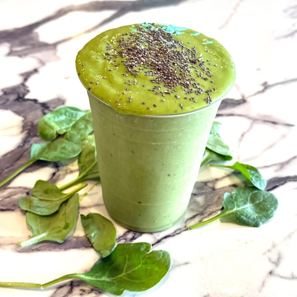 Green smoothie in a clear cup, topped with chia seeds, surrounded by spinach leaves on marble.