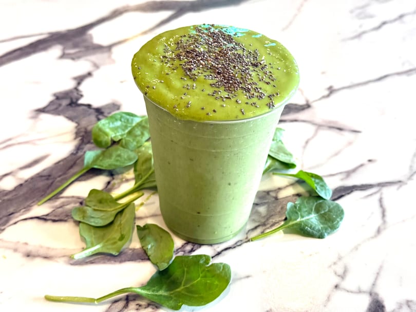 Green smoothie in a clear cup, topped with chia seeds, surrounded by spinach leaves on marble.