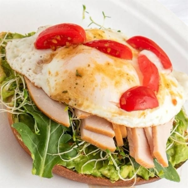 Open-faced sandwich with avocado, turkey, fried egg, cherry tomatoes, and sprouts on a white plate.